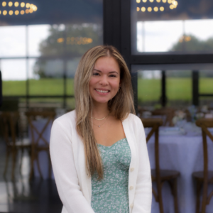Smiling young woman with long blond hair wearing a light cardigan and teal floral dress, indoors at a dining venue with tables in the background